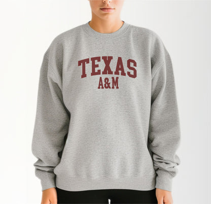 A model wears the Sport Grey Adult Unisex Texas A&M Varsity Crewneck Sweatshirt.  The ﻿Texas A&M Varsity﻿ graphic is in bold Maroon in a Collegiate style.