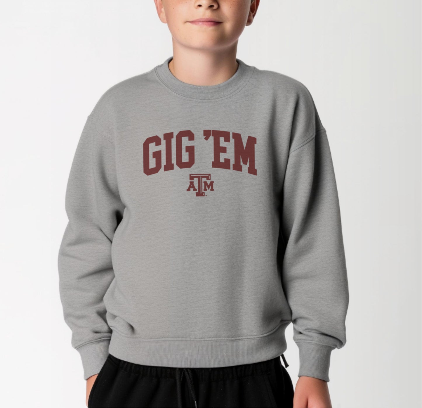 A model wears the Sport Grey Youth Unisex Texas A&M Gig 'Em Collegiate Crewneck Sweatshirt.  The ﻿Texas A&M Gig 'Em Collegiate﻿ graphic is in bold Maroon in a Collegiate style.