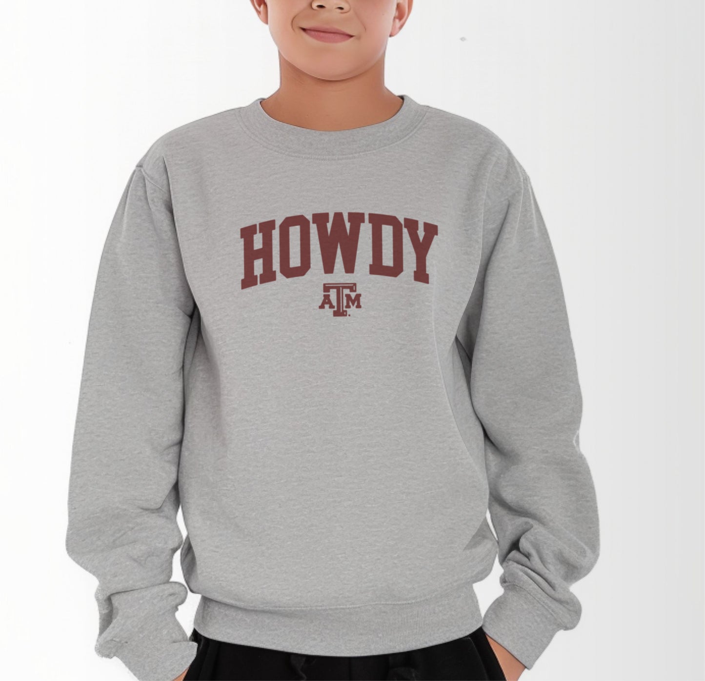 A model wears the Sport Grey Youth Unisex Texas A&M Howdy Varsity Crewneck Sweatshirt.  The ﻿Texas A&M Howdy Varsity﻿ graphic is in bold Maroon in a Collegiate style.