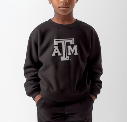 A model wears the Black Youth Unisex Texas A&M Logo Crewneck Sweatshirt.  The ﻿Texas A&M Logo﻿ graphic is in bold White in a Varsity style.