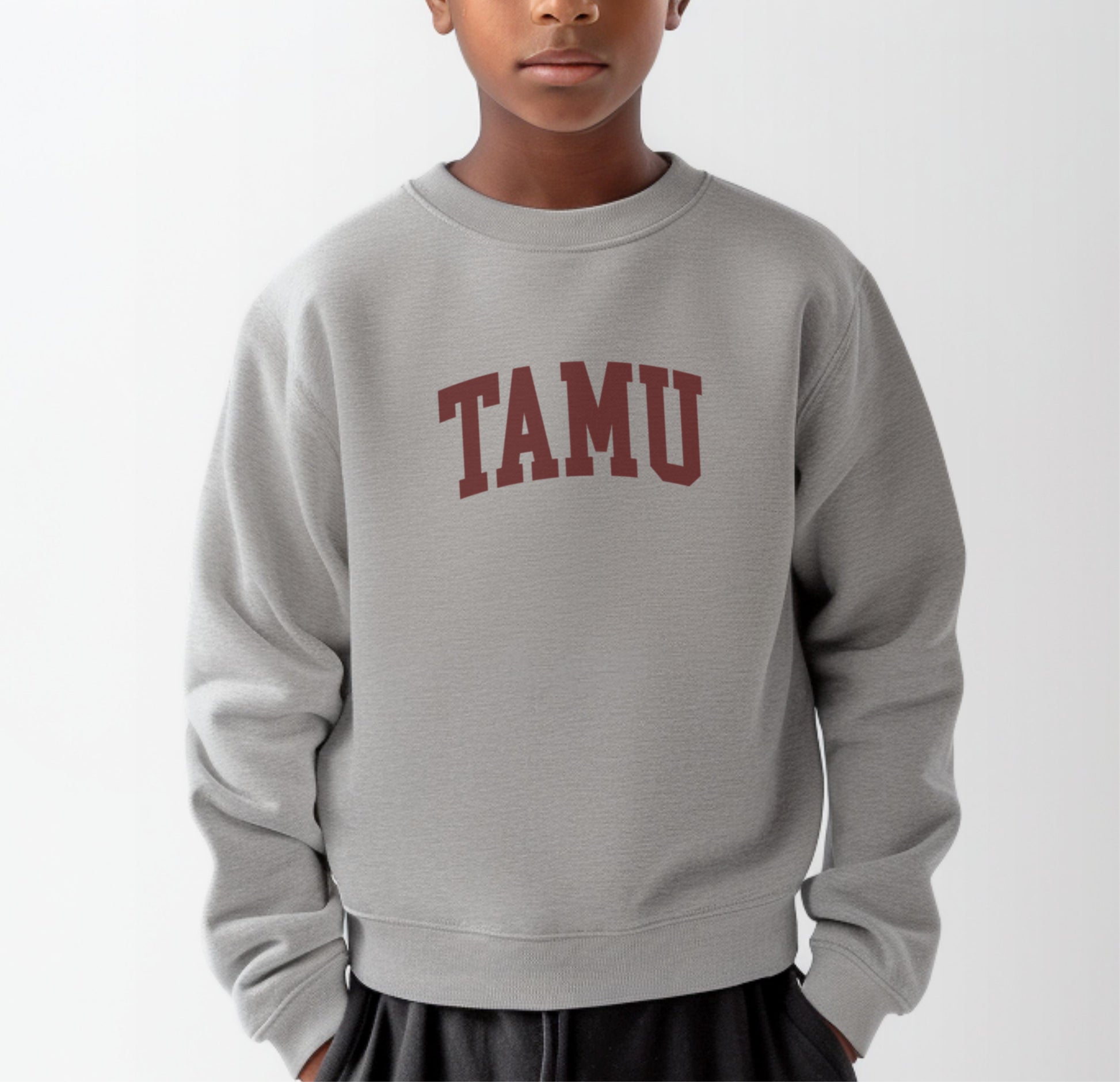 A model wears the Sport Grey Youth Unisex Texas A&M TAMU Crewneck Sweatshirt.  The ﻿Texas A&M TAMU﻿ graphic is in bold Maroon in a Collegiate style.