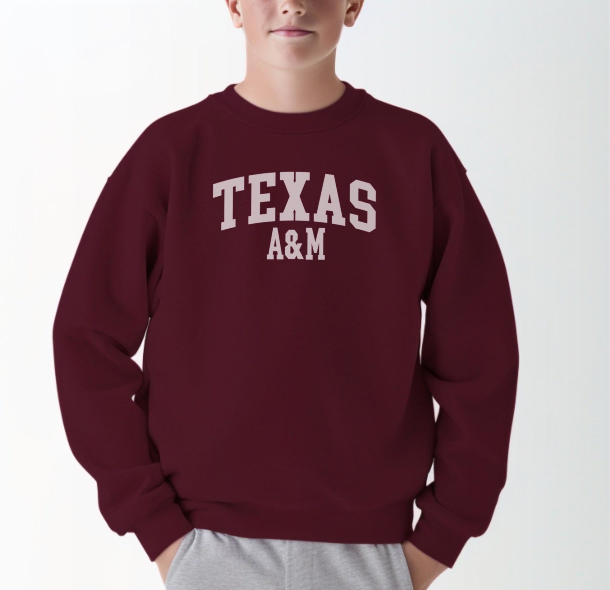 A model wears the Maroon Youth Unisex Texas A&M Varsity Crewneck Sweatshirt.  The ﻿Texas A&M Varsity﻿ graphic is in bold White in a Collegiate style.