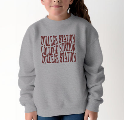 A model wears the Sport Grey Youth Unisex College Station Western Crewneck Sweatshirt.  The ﻿College Station Western﻿ graphic is in bold Maroon in a Western style.