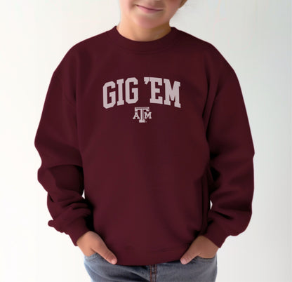 A model wears the Maroon Youth Unisex Texas A&M Gig 'Em Collegiate Crewneck Sweatshirt.  The ﻿Texas A&M Gig 'Em Collegiate﻿ graphic is in bold White in a Collegiate style.