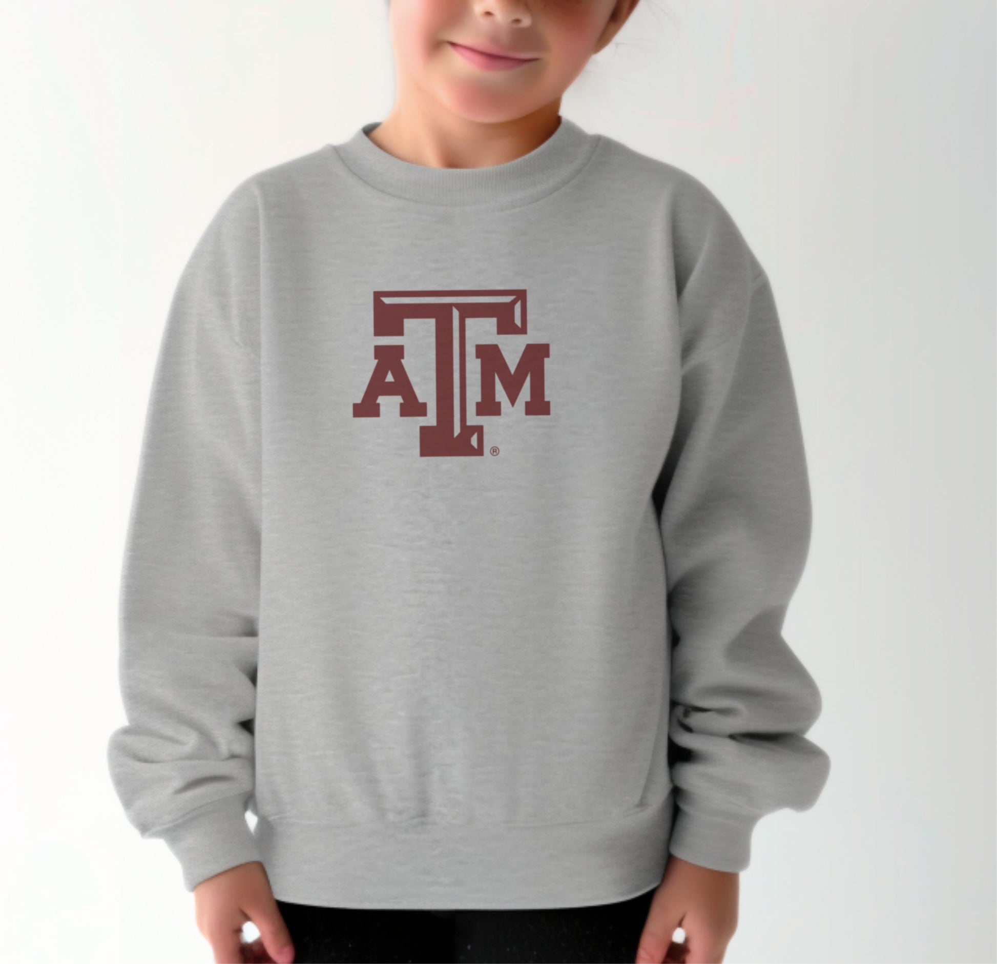 A model wears the Sport Grey Youth Unisex Texas A&M Logo Crewneck Sweatshirt.  The ﻿Texas A&M Logo﻿ graphic is in bold Maroon in a Varsity style.