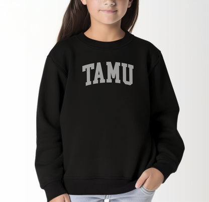 A model wears the Black Youth Unisex Texas A&M TAMU Crewneck Sweatshirt.  The ﻿Texas A&M TAMU﻿ graphic is in bold White in a Collegiate style.