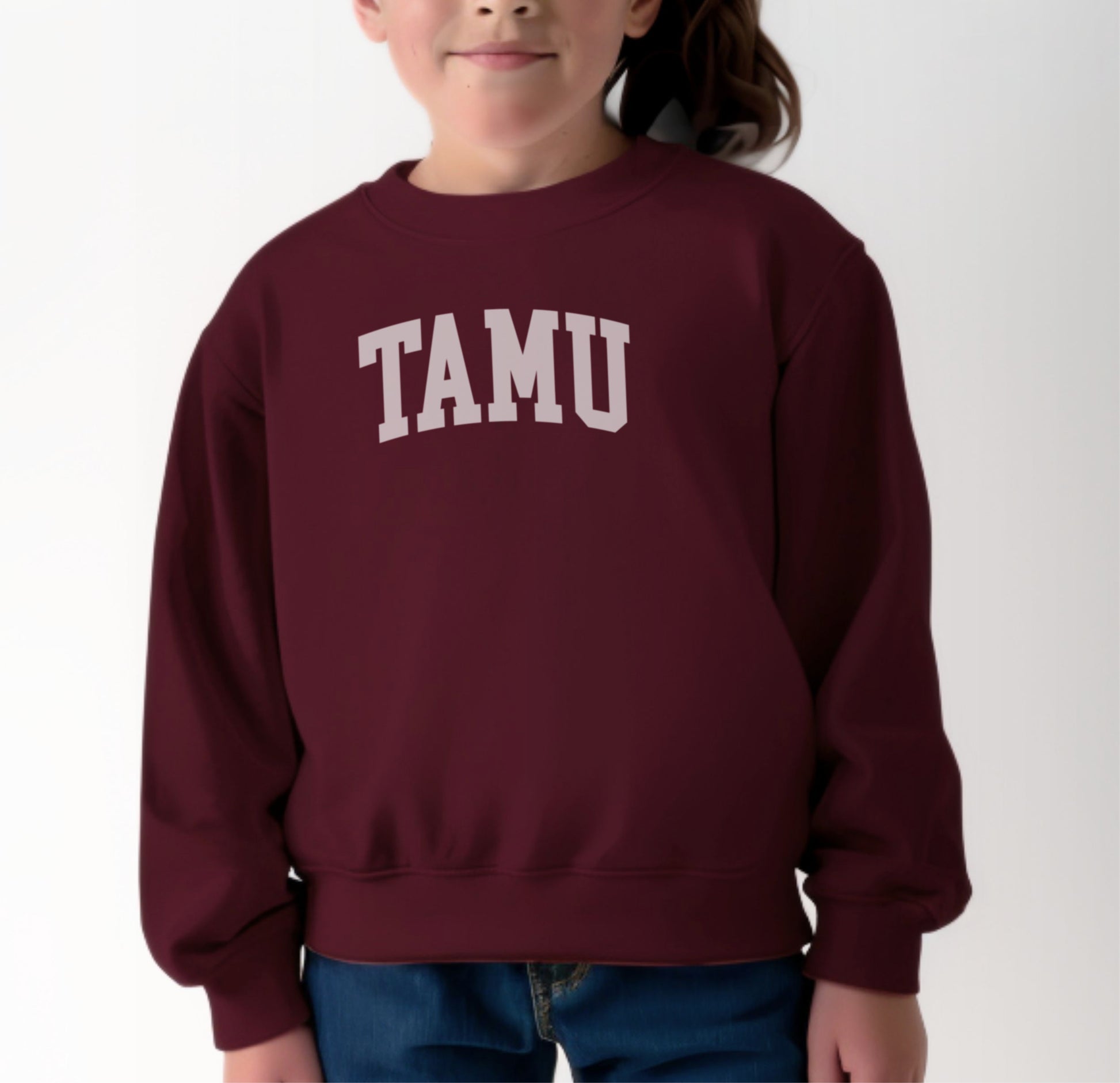 A model wears the Maroon Youth Unisex Texas A&M TAMU Crewneck Sweatshirt.  The ﻿Texas A&M TAMU﻿ graphic is in bold White in a Collegiate style.