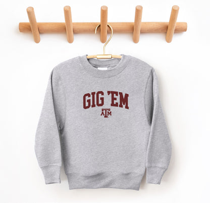 The Sport Grey Youth Unisex Texas A&M Gig 'Em Collegiate Crewneck Sweatshirt lays flat on a white background. The ﻿Texas A&M Gig 'Em Collegiate﻿ graphic is in bold Maroon in a Collegiate style.