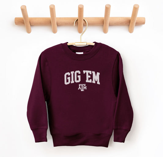 The Maroon Youth Unisex Texas A&M Gig 'Em Collegiate Crewneck Sweatshirt lays flat on a white background. The ﻿Texas A&M Gig 'Em Collegiate﻿ graphic is in bold White in a Collegiate style.