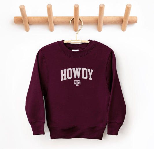 The Maroon Youth Unisex Texas A&M Howdy Varsity Crewneck Sweatshirt lays flat on a white background. The ﻿Texas A&M Howdy Varsity﻿ graphic is in bold White in a Collegiate style.