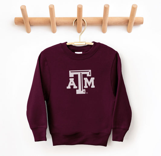 The Maroon Youth Unisex Texas A&M Logo Crewneck Sweatshirt lays flat on a white background. The ﻿Texas A&M Logo﻿ graphic is in bold White in a Varsity style.