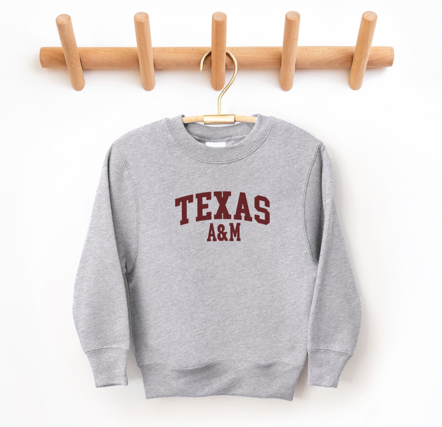 The Sport Grey Youth Unisex Texas A&M Varsity Crewneck Sweatshirt lays flat on a white background. The ﻿Texas A&M Varsity﻿ graphic is in bold Maroon in a Collegiate style.