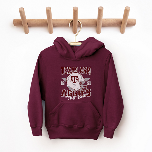 A model wears the Maroon Texas A&M Aggies Football Hooded Sweatshirt.  The ﻿Texas A&M Aggies Football﻿ graphic is in bold Multicolor in a Varsity style.